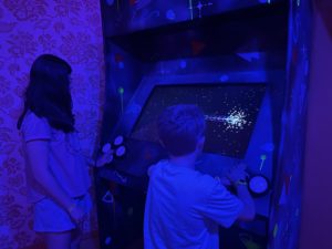 Two kids playing a video game arcade machine in a blue lit room enjoying interactive family fun at Factory Obscura.